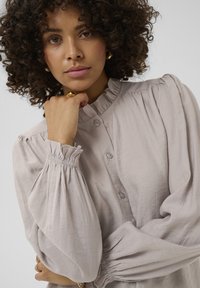 Lys grå bluse med en høj, rynket krave, knaplukning fortil og pufærmer. Blødt, struktureret stof med sammenkrøllede manchetter.