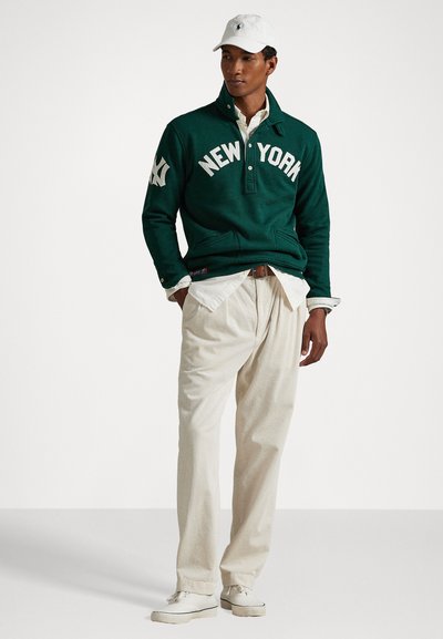 Zelený pulóver s nápisom "NEW YORK", v kombinácii s béžovými kordovým nohavicami a bielymi teniskami. Biely klobúk dopĺňa outfit.
