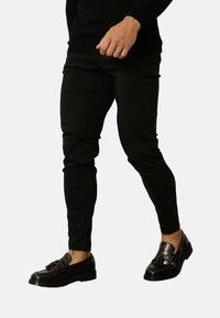 Pantalons noirs slim-fit en tissu extensible, présentant une texture lisse et un design fuselé. Portés avec des mocassins noirs.