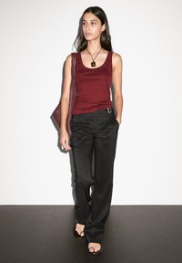 Débardeur bordeaux, pantalon large noir, portant un sac à bandoulière en osier et des sandales marron. Tissus lisses et style décontracté.