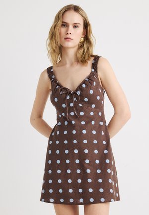 Νεαρή γυναίκα που φοράει καφέ αμάνικο φόρεμα με ανοιχτό μπλε πουά και λεπτομέρεια δέσιμου στο μπροστινό μέρος, στέκεται μπροστά σε απλό φωτεινό φόντο.