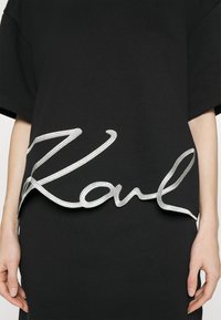 Černé tričko s velkým stříbrným vyšitým nápisem "Karl" u lemu. Krátké rukávy, volný střih, hladká bavlněná textura.