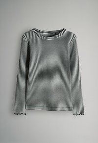 Svartvit randig långärmad skjorta med rund halsringning och en liten volangdetalj vid ärmsluten, hängande mot en grå bakgrund.