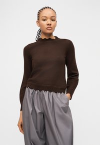 Νεαρό άτομο με πλεγμένα μαλλιά, που φοράει σκούρα καφέ μακρυμάνικη μπλούζα και άνετο ανοιχτό γκρι παντελόνι, στέκεται με το ένα χέρι στην τσέπη.