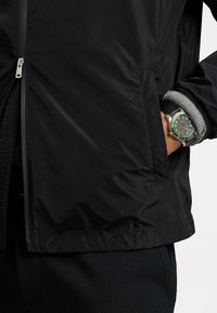 Musta vetoketjullinen takki, jossa on sileä pinta, hopeiset metalliosat ja sivutasku. Ranteessa näkyy vihreällä kellotaululla varustettu kello, jossa on musta hihna.