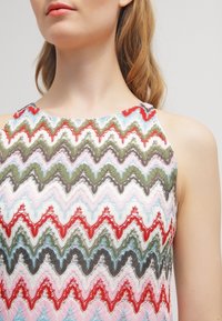 Robe colorée à motifs en zigzag, présentant des nuances de rouge, vert, bleu et rose. La texture du tissu est détaillée avec des éléments en dentelle intriqués.