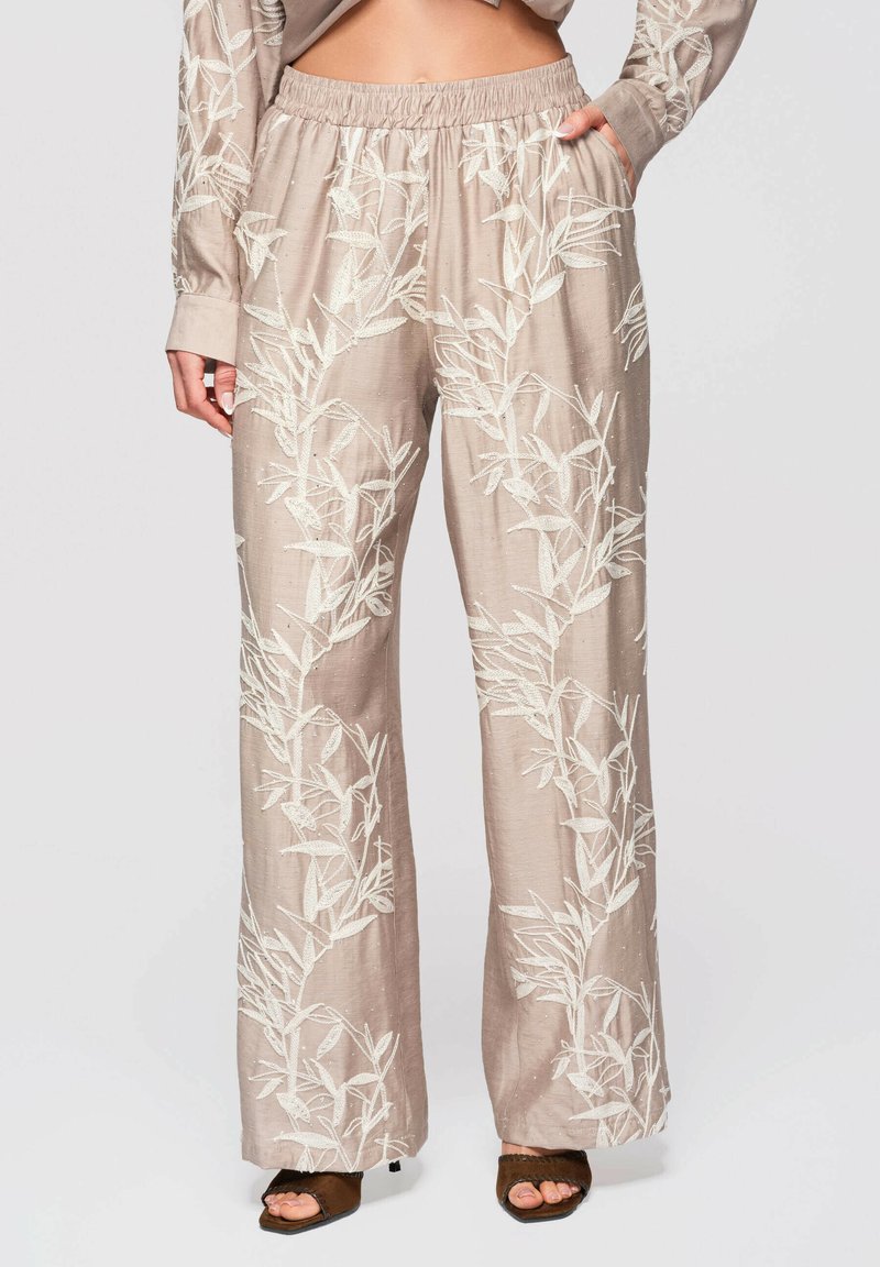 Pantalon beige à jambes larges avec broderie florale blanche, taille élastique et poches latérales, porté avec des talons ouverts marron.