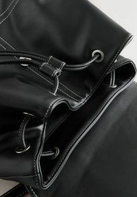 Sac en cuir noir avec fermeture à cordon, présentant des surpiqûres blanches contrastées et des œillets métalliques. Texture lisse avec un design minimaliste.