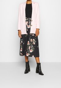 Světle růžové sako se zlatými knoflíky přes černé květinové midi šaty s růžovými a bílými květy. Černé kotníkové boty dotvářejí celkový dojem.