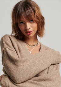 Suéter oversized beige con escote en V, textura suave y mangas sueltas. Collares de plata en capas con diseños variados y un colgante en forma de corazón.