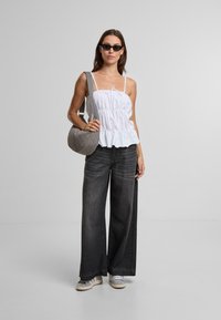 Biela peplum blúzka s tenkými ramienkami, šedé široké nohavice, šedá prešívaná ramenná taška a biele tenisky, na jednofarebnom pozadí.