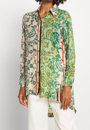 Camisa de manga larga con botones y patrones mixtos de flores paisley en verde y beige, combinada con pantalones blancos.