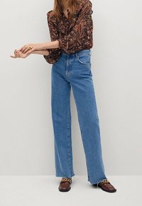 Chemisier à manches longues à motifs floraux, boutonné à l'avant, associé à un jean bleu taille haute à jambes droites et à des mocassins marron ornés de chaînes dorées.