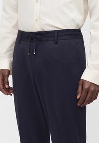 Homme portant un pantalon bleu marine avec une taille à cordon et une chemise beige clair à manches longues boutonnée.