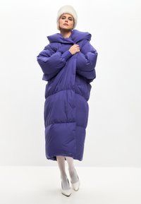 Palton pufos violet cu glugă supradimensionată și design matlasat voluminos. Material neted și reflectant, lungime până la genunchi, asortat cu tocuri albe.