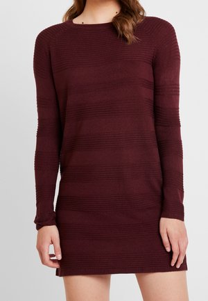 Femme portant une robe en maille bordeaux à manches longues avec de subtiles rayures horizontales texturées, debout devant un fond gris uni.