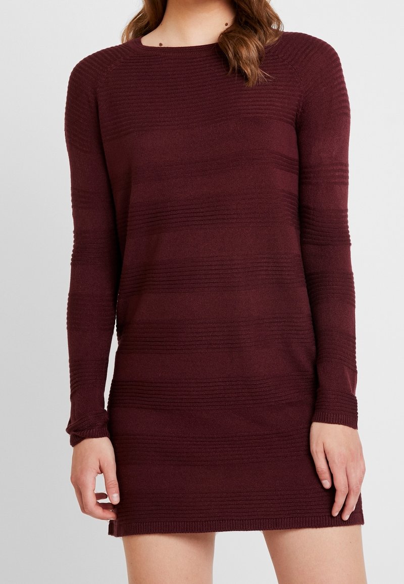 Femme portant une robe en maille bordeaux à manches longues avec de subtiles rayures horizontales texturées, debout devant un fond gris uni.
