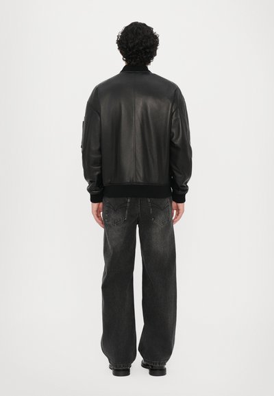 Black leather bomber jacket with ribbed cuffs and hem, worn over dark grey, loose-fitting jeans. Shot from the back against a white background.