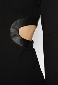 Vêtement noir avec une découpe courbée sur le côté, bordée d'une finition en cuir noir texturé, exposant la peau en dessous.
