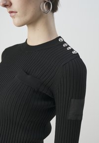 Svart ribbat linne med rundad halsringning, horisontell textur, silverfärgade knappar på axeln och en liten ficka fram.