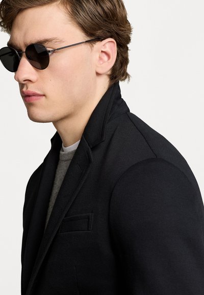 Blazer noir avec un col cranté et une poche poitrine simple, porté sur un pull gris. Le mannequin porte des lunettes de soleil rondes et sombres.