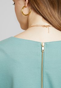 Ljus turkos topp med en framträdande gyllene dragkedja baktill och ett slätt, texturerat tyg. Kedjehalsbandssmycke synligt vid nackens bas.