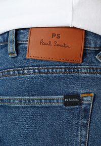 Blå denimjeans med brun läderlapp märkt "PS Paul Smith." Har gula sömmar och ett litet svart etikett.