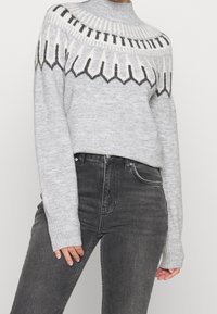 Suéter gris de punto con cuello alto, que presenta un patrón geométrico en blanco y negro a lo largo de la parte superior, combinado con jeans negros de cintura alta.