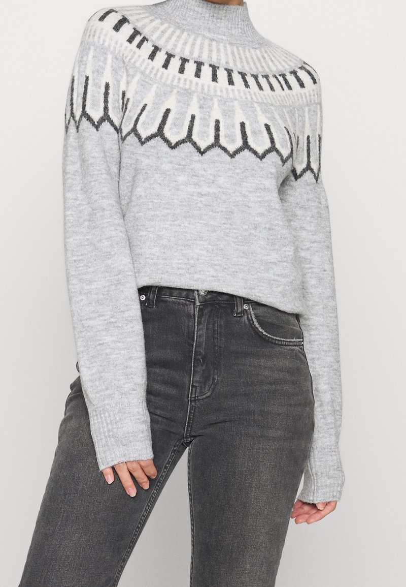 Suéter gris de punto con cuello alto, que presenta un patrón geométrico en blanco y negro a lo largo de la parte superior, combinado con jeans negros de cintura alta.
