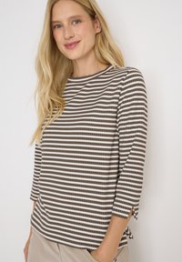 Femme blonde portant une chemise à rayures marron et blanche à manches longues avec un pantalon beige, souriant légèrement contre un fond blanc uni.