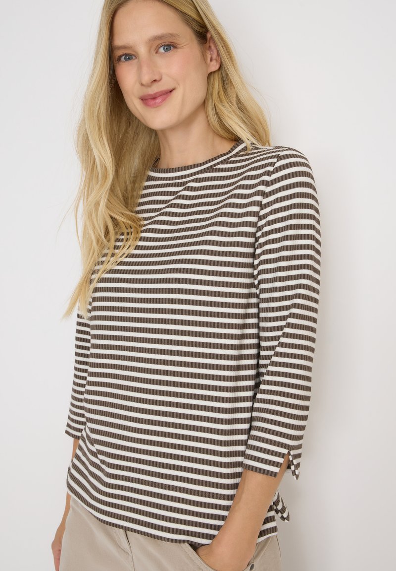 Femme blonde portant une chemise à rayures marron et blanche à manches longues avec un pantalon beige, souriant légèrement contre un fond blanc uni.