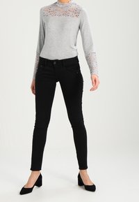 Grå långärmad topp med spetsdetaljer på axlarna och bröstet, tillsammans med figursydda svarta jeans och svarta spetsiga klackar.