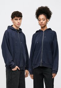 Navy zip-up hoodies made from soft fabric feature a relaxed fit, front pockets, drawstrings, and ribbed cuffs, paired with dark jeans.