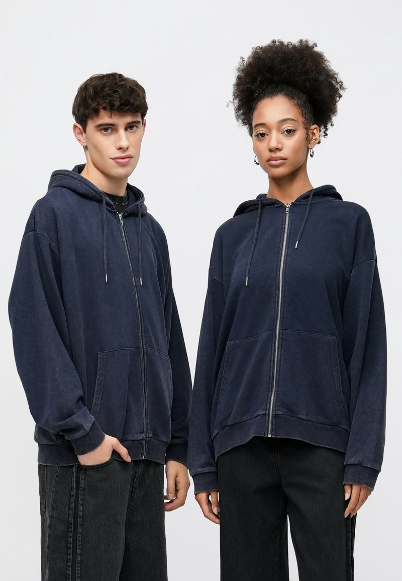 Navy zip-up hoodies made from soft fabric feature a relaxed fit, front pockets, drawstrings, and ribbed cuffs, paired with dark jeans.
