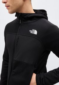 Muž nosící černou zipovou mikinu s kapucí a logem "The North Face" na hrudi, s rukou opřenou o bok, otočený vlevo.