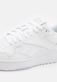 Sneaker de couro branco com sola de borracha texturizada, apresentando perfurações, atacadores planos e detalhes de marca discretos. Forma suave e arredondada.