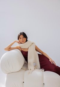 Crèmekleurige wollen sjaal met franjes aan de randen, gedrapeerd over een model dat een bordeauxrode satijnen jurk draagt, zittend op een witte pluchen fauteuil.