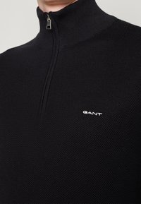 Pull noir en maille avec col demi-fermeture éclair et petit logo blanc "GANT" brodé sur la partie supérieure de la poitrine.