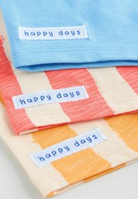 Trzy bawełnowe koszulki T-shirt: jasnoniebieska, brzoskwiniowa w czerwone paski oraz kremowa w pomarańczowe paski. Każda ma niebieską metkę z napisem "happy days".
