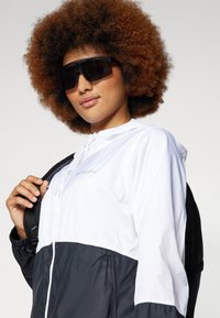 Weißer und marineblauer Windbreaker mit Kordelzugkapuze, mit einem Reißverschluss vorn, elastischen Bündchen und Logo-Detail. Das Modell trägt übergroße schwarze Sonnenbrillen.