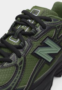 Κοντινή λήψη αθλητικού παπουτσιού σε πράσινο και μαύρο χρώμα με υφή από πλέγμα, μαύρα κορδόνια και το ανεβασμένο πράσινο λογότυπο "N" στην πλαϊνή επιφάνεια.