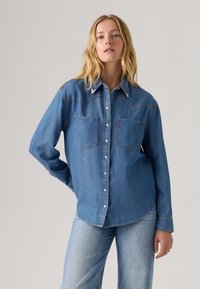 Denim-Hemd mit Druckknöpfen, zwei Brusttaschen, spitzem Schulterdesign, mittlerem Blauton und sichtbaren Nähten. Weicher, leichter Stoff.
