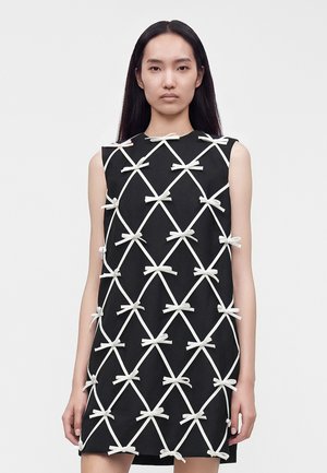 Frau mit langen dunklen Haaren, die ein ärmelloses schwarzes Kleid trägt, das mit weißen Schleifen in einem Rautenmuster verziert ist, steht vor einem schlichten Hintergrund.