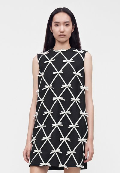Femme aux longs cheveux foncés portant une robe noire sans manches décorée de nœuds de ruban blanc en motif losange, se tenant devant un fond uni.