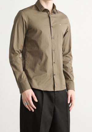 Homme portant une chemise cintrée vert olive à boutons avec manches longues et un pantalon noir, debout les mains détendues le long du corps.