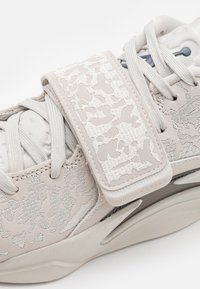 Bézs színű sportcipő texturált velúr és szövet részletekkel. Jellemző egy feltűnő pánt fehér dombornyomott mintával és szürke fűzőkkel. Lekerekített talp kialakítás.