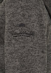 Šedý pletený svetr s texturovaným vzorem. Obsahuje jemně vyšitý logo deštníku na rukávu.