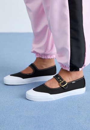 Stopy w czarno-białych butach Vans w stylu Mary Jane z paskami na klamrę, w połączeniu z różowo-czarnymi dresami dwukolorowymi na niebieskim dywanie.