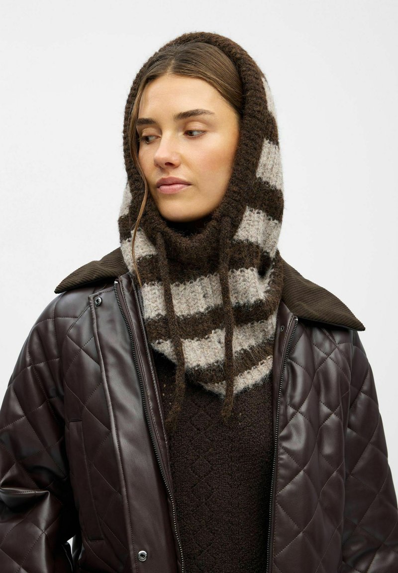 Brun quiltet jakke over en mørk sweater, prydet med et stribet uldscarf i brun og grå med to hængende bånd.