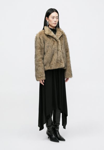 Ψεύτικη γούνινη καφέ ζακέτα με γιακά, φορεμένη πάνω από ένα μακρύ μαύρο φόρεμα. Slim μαύρες μπότες ολοκληρώνουν την εμφάνιση. Η υφή είναι μαλακή και αφράτη.
